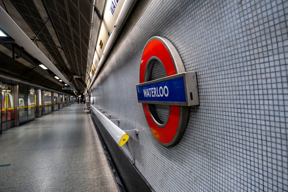 A wide, empty platform at Waterloo underground station, London, featuring a tiled wall with the roundel London Underground sign displaying 'Waterloo' in white text on a blue background. The platform has a smooth, grey terrazzo floor and is lined with glass barriers separating passengers from the train tracks. In the background, there are a few travelers waiting, with some standing close to the barriers and others further away. Overhead, the station's ceiling is dark with visible structural elements and lighting fixtures providing illumination. A yellow safety strip runs along the edge of the platform for passenger safety. This station interior is typical for London Underground, with clean, modern finishes and functional signage, supporting safe and efficient home relocation and furniture transport processes for those moving near Colliers Wood and other South London locations. Occasionally, such images evoke the logistical planning involved in moving and packing belongings for effective transport during house removals.