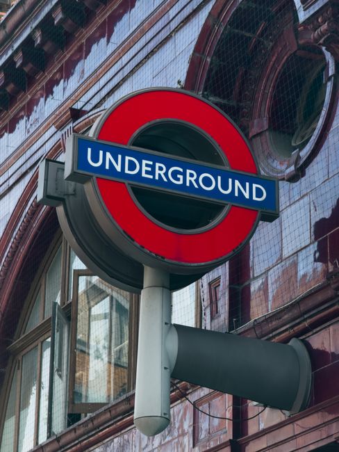 A close-up view of the London Underground roundel sign with a red circle and blue bar displaying the word 'UNDERGROUND' in white lettering, mounted on a white pole attached to a building with reflective glass windows and brickwork. The background includes the urban exterior of a city street with part of the building facade visible, and natural daylight illuminates the scene. This image depicts typical surroundings for London transport signage, often captured during house and furniture removals to highlight urban moving logistics and the environment adjacent to residential areas near Colliers Wood tube station. Man and a Van Colliers Wood occasionally use such landmarks to contextualise home relocation and furniture transport services, though no moving process is visible in this specific image.