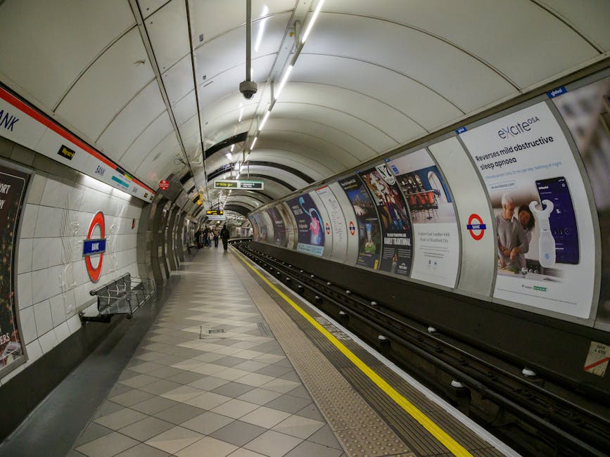 A wide, empty platform at Waterloo underground station, London, featuring a tiled wall with the roundel London Underground sign displaying 'Waterloo' in white text on a blue background. The platform has a smooth, grey terrazzo floor and is lined with glass barriers separating passengers from the train tracks. In the background, there are a few travelers waiting, with some standing close to the barriers and others further away. Overhead, the station's ceiling is dark with visible structural elements and lighting fixtures providing illumination. A yellow safety strip runs along the edge of the platform for passenger safety. This station interior is typical for London Underground, with clean, modern finishes and functional signage, supporting safe and efficient home relocation and furniture transport processes for those moving near Colliers Wood and other South London locations. Occasionally, such images evoke the logistical planning involved in moving and packing belongings for effective transport during house removals.