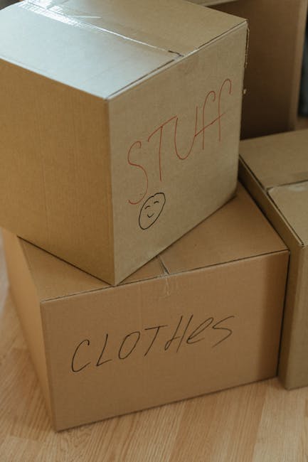 A close-up view of two cardboard boxes placed on a wooden floor inside a property, likely during a home relocation. The top box has the word 'STUFF' handwritten in red marker on its side, along with a simple smiley face. The lower box is labeled 'CLOTHES' in the same handwriting style. Both boxes appear to be sealed and are made of standard brown cardboard. Additional boxes are partially visible nearby, indicating an ongoing packing or moving process. The scene reflects typical packing materials used during furniture transport and packing and moving activities, with the boxes arranged for easy lifting and carrying, supported by the presence of other packing items. This setup aligns with professional removals services like those offered by Man and a Van Colliers Wood, focusing on efficient home relocation and packing logistics.