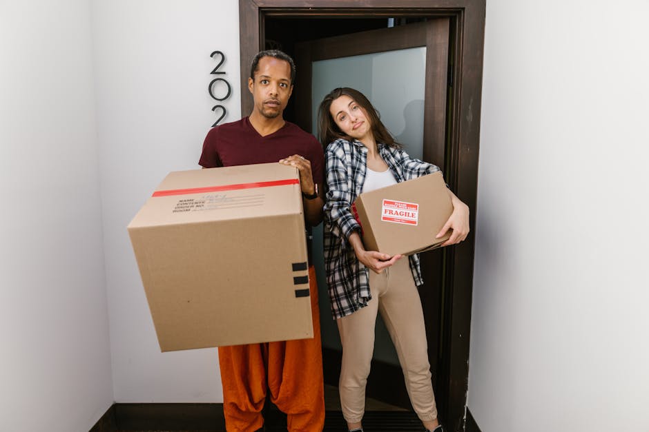 A man and a woman stand inside a residence near a doorway, each holding a cardboard box as part of the home relocation process. The man, wearing a maroon top and orange trousers, holds a large box in front of him, while the woman, dressed in a checkered shirt and beige pants, cradles a smaller box labeled 'Fragile' close to her chest. The interior features plain white walls, and the doorway has a dark wooden frame, likely leading to another room or the exterior. The lighting appears natural, illuminating the scene clearly. The boxes are made of cardboard, some reinforced with packing tape, and the woman’s box has bubble wrap or packing material visible at the edges. The process depicted suggests careful handling typical of furniture transport and packing for moving services, aligning with professional removals by Man and a Van Colliers Wood, who manage loading and unloading procedures at residential properties.